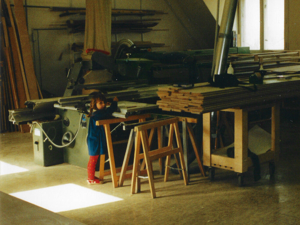 Ansicht einer Holzwerkstatt mit Werkbänken, Holzplatten und Maschinen; im Vordergrund steht ein Kind, das an einem Arbeitstisch lehnt.