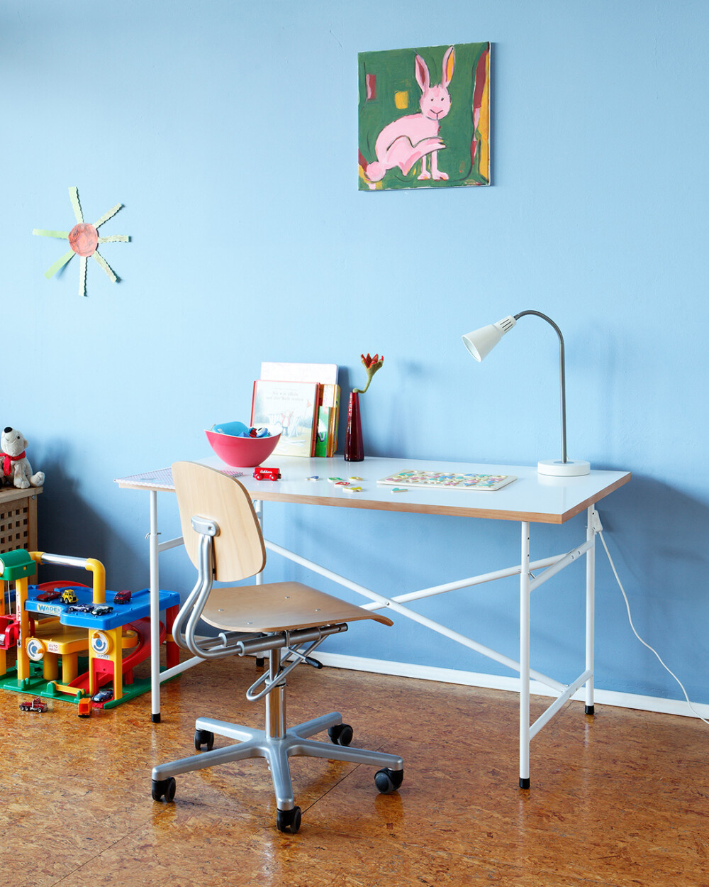 E2 Kinder-Schreibtisch mit weißer Platte im Kinderzimmer, genutzt als Arbeitsplatz mit Lampe, Büchern und Spielzeug.